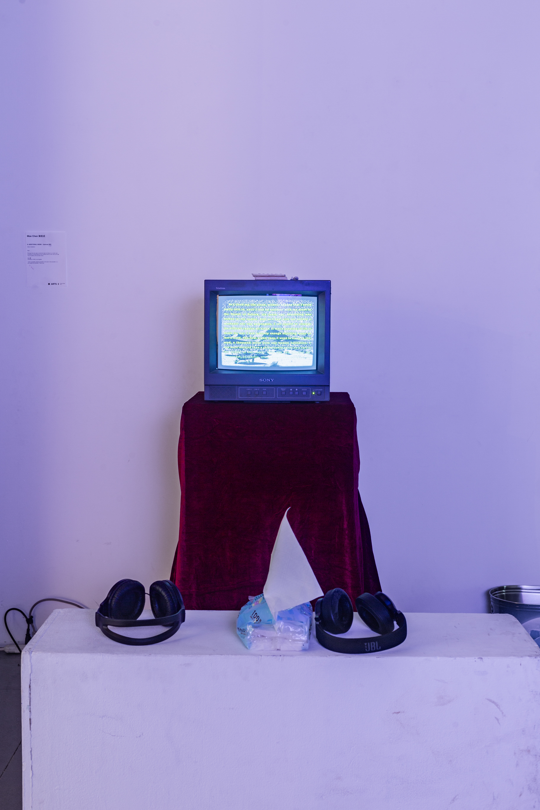 A CRT tv with artistic visuals on it, ontop of a podium covered in a table cloth in a white room. In front of it is seating with two headphones.